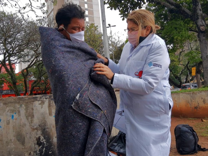 Profissional do Consultório na Rua ampara uma pessoa em situação de rua numa praça, que está enrolada a um cobertor
